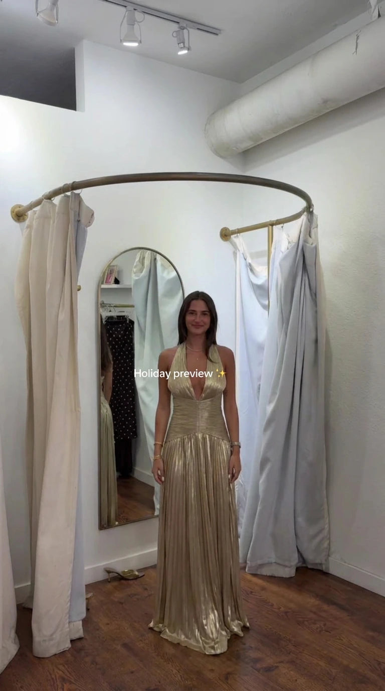 Front full-length view of model wearing gold metallic pleated halter maxi gown in fitting room